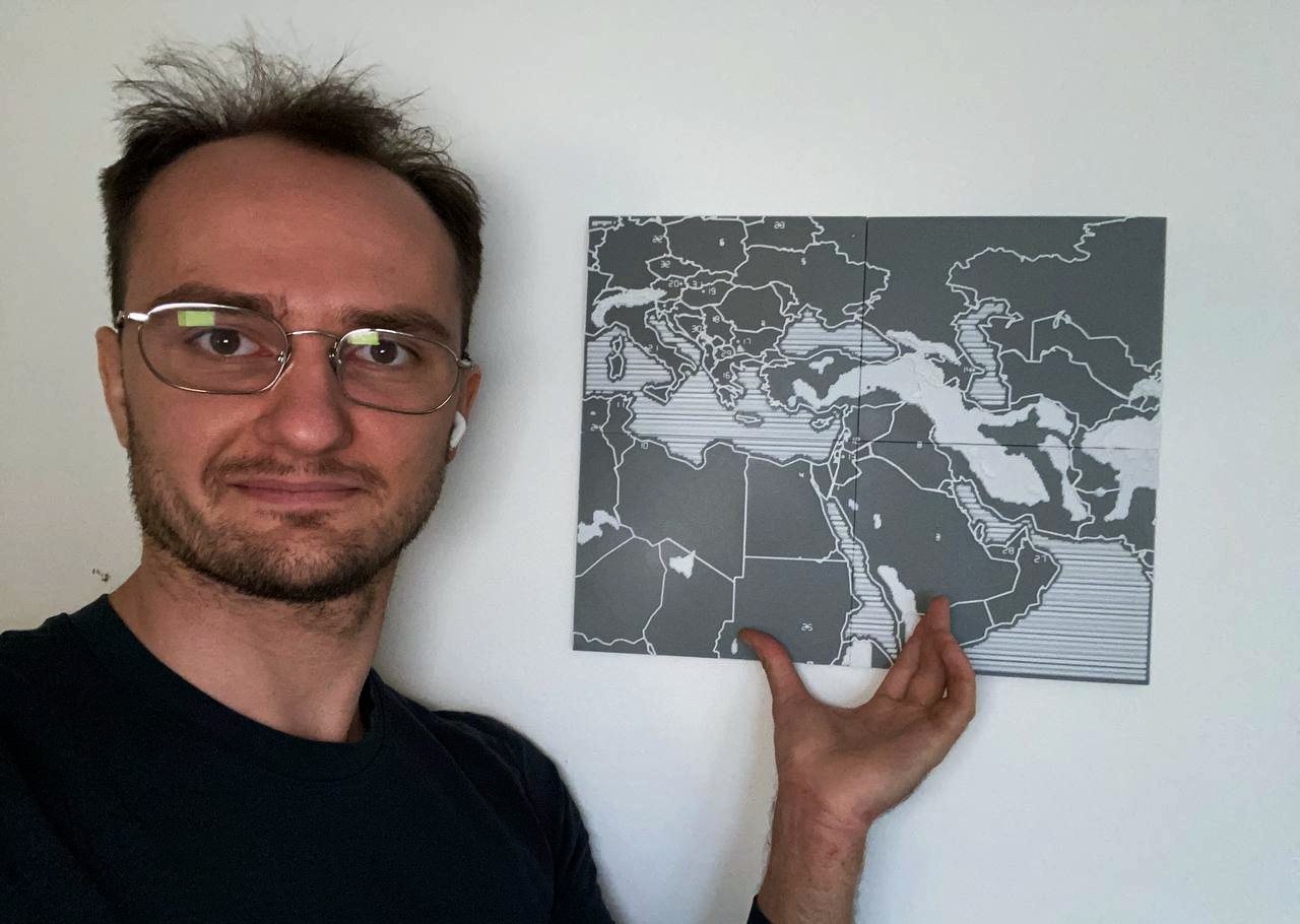 Gleb standing beside the assembled tactile map mounted on a wall, one hand resting against it.