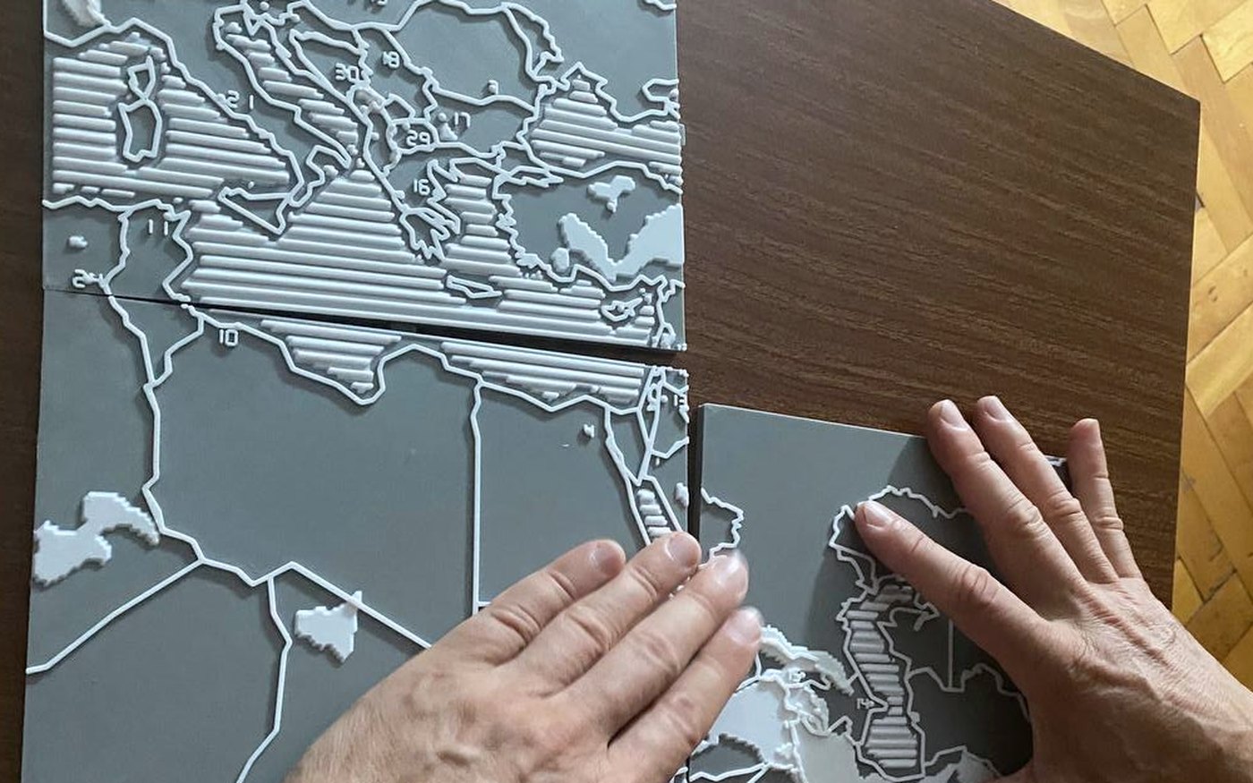 Close photograph of two printed tiles side by side on a dark table, with a hand resting on one of them: the borders stand up as white ridges, the seas are ribbed with parallel lines and the mountains rise in flat steps.