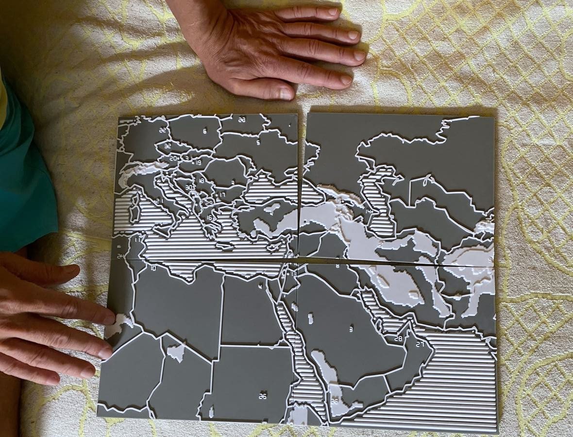 Two hands resting on the assembled tactile map, seen from above. Four printed tiles form one map of Europe, the Middle East and North Africa, with raised white borders, ribbed seas and small raised numbers inside the larger countries.