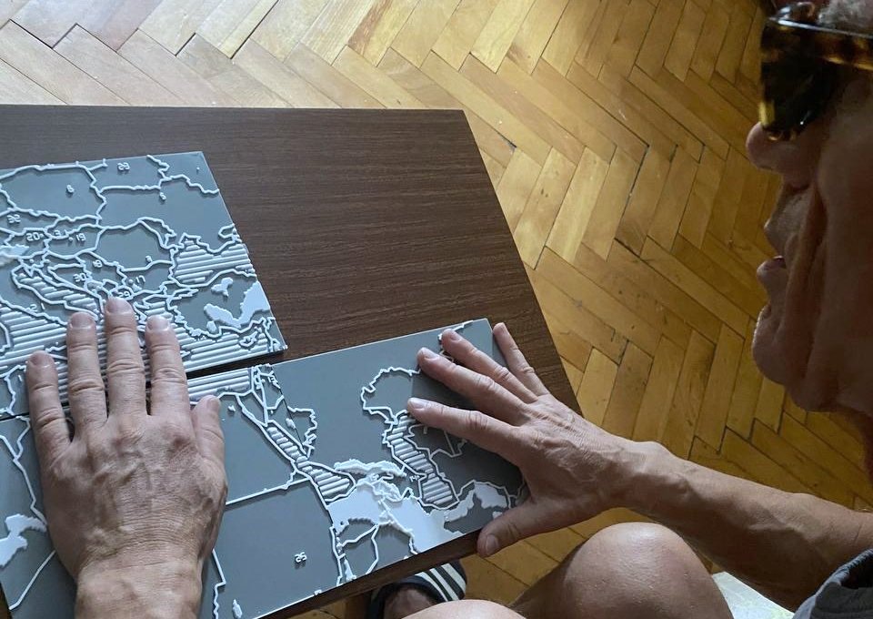 A blind man in dark glasses leans over a table with both hands flat on two tiles of the tactile map, reading the raised borders with his fingers.